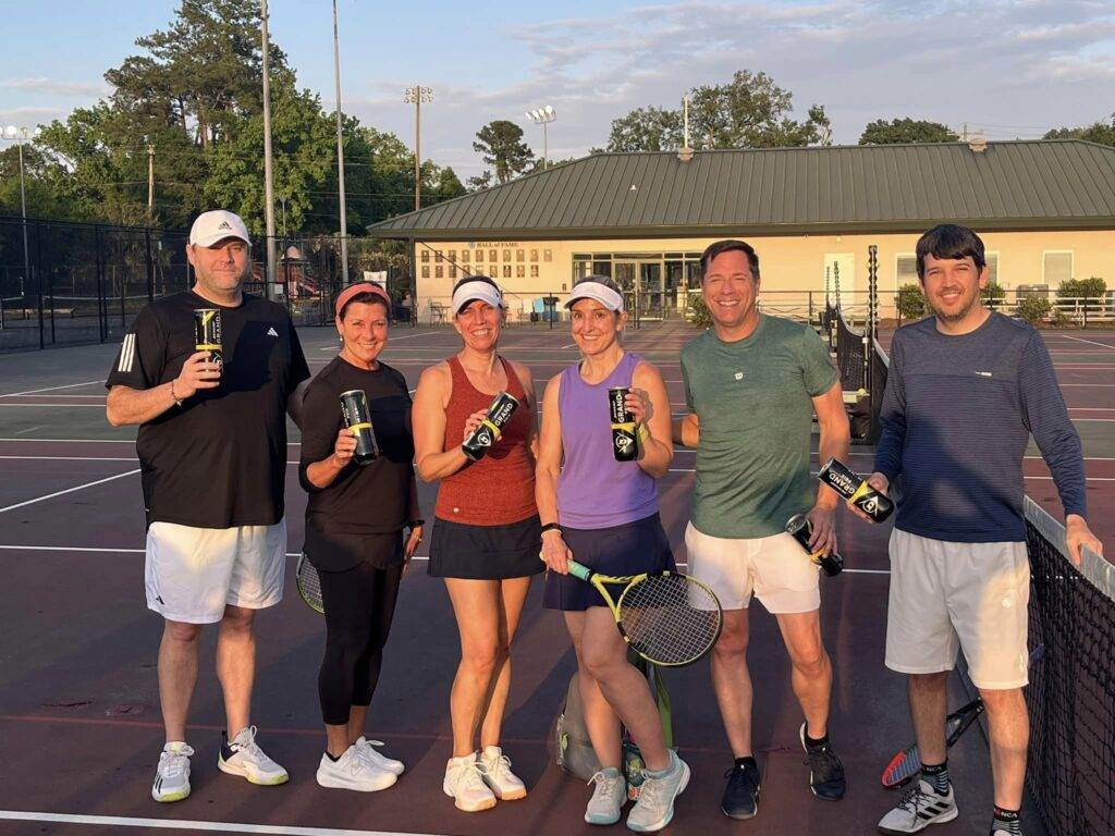 A group of smiling LowCountry Tennis Players on a tennis court enjoying some gifts from National Tennis Month.