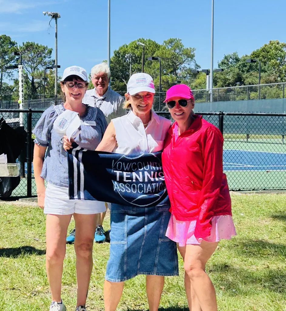 Four smiling adults pose outdoors in front of tennis courts, holding a “LowCountry Tennis Association” banner. Three women wear hats and light clothing; a man stands behind them. It is sunny with green trees in the background.