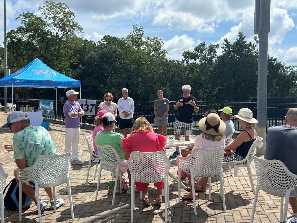 A group of people sit on white chairs around tables outdoors at an LCTA event, while several others stand and speak in front of them. A blue canopy tent and trees are in the background beneath a partly cloudy sky.