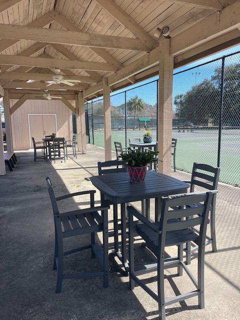 Covered patio with gray tables and chairs overlooks tennis courts through a chain-link fence. A small potted plant sits on a table in the foreground. Sunlight casts shadows on the concrete floor.