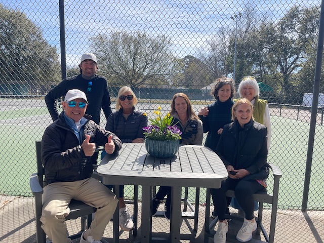 Seven adults pose and smile around a table with a flower pot on it, outdoors by a tennis court on a sunny day. They are dressed in jackets, and trees are visible in the background.