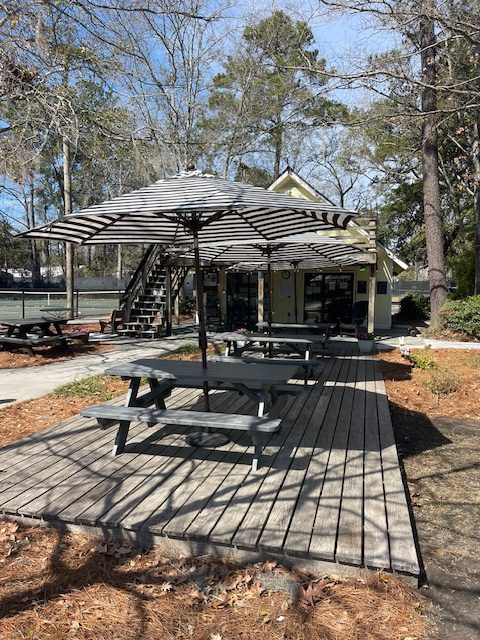 Gray picnic tables with black and white striped umbrellas sit on a wooden deck near a small building, surrounded by trees and tennis courts on a sunny day.
