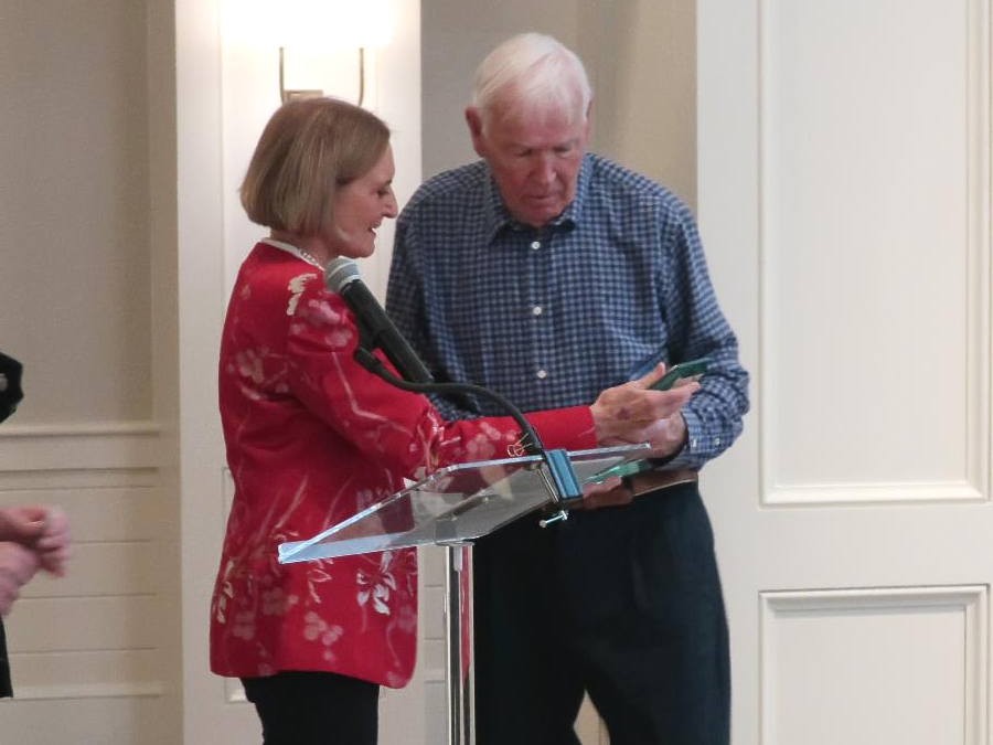 A woman in a red floral jacket hands an award or plaque to an older man in a blue checkered shirt as they stand together at a podium with a microphone.