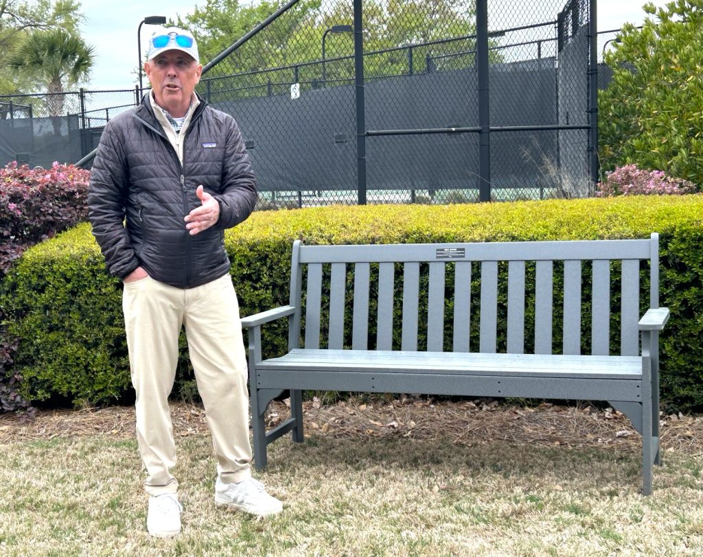 A man in a black jacket, white pants, and a cap stands next to an empty gray bench on a grassy area, with green bushes and a tall fence in the background.
