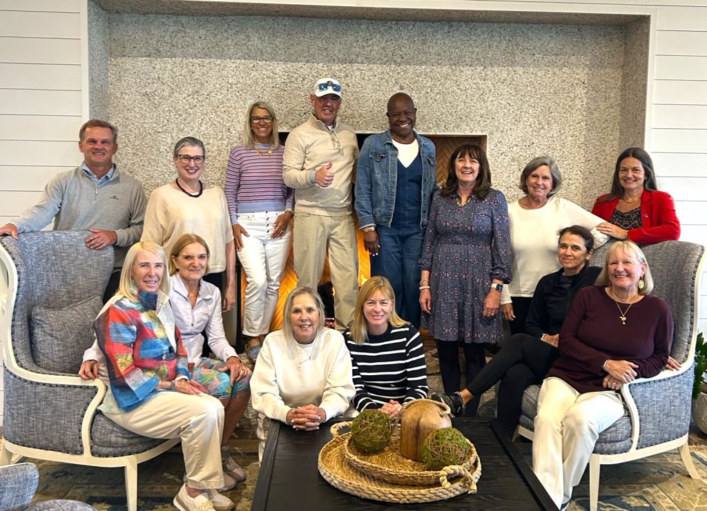 A group of fourteen adults, both men and women, pose together indoors in a living room setting. Some are seated on armchairs and a sofa, while others stand behind them, all smiling at the camera.