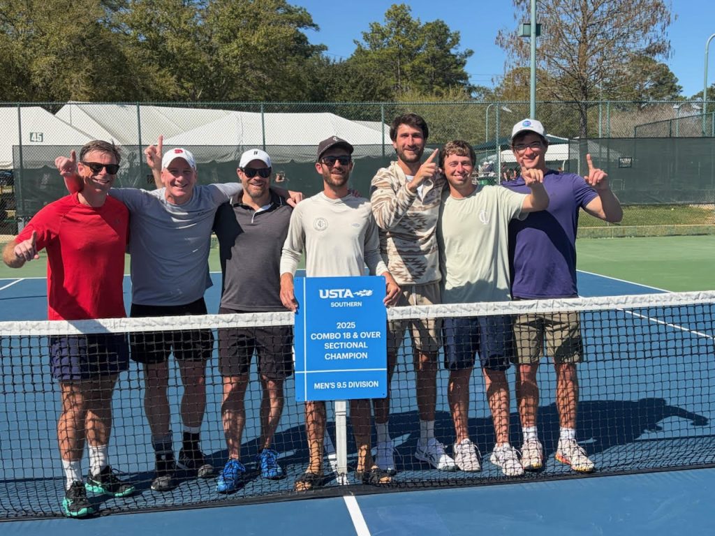 Seven men stand on a tennis court behind a net, smiling, some holding up one finger. They hold a blue sign that reads