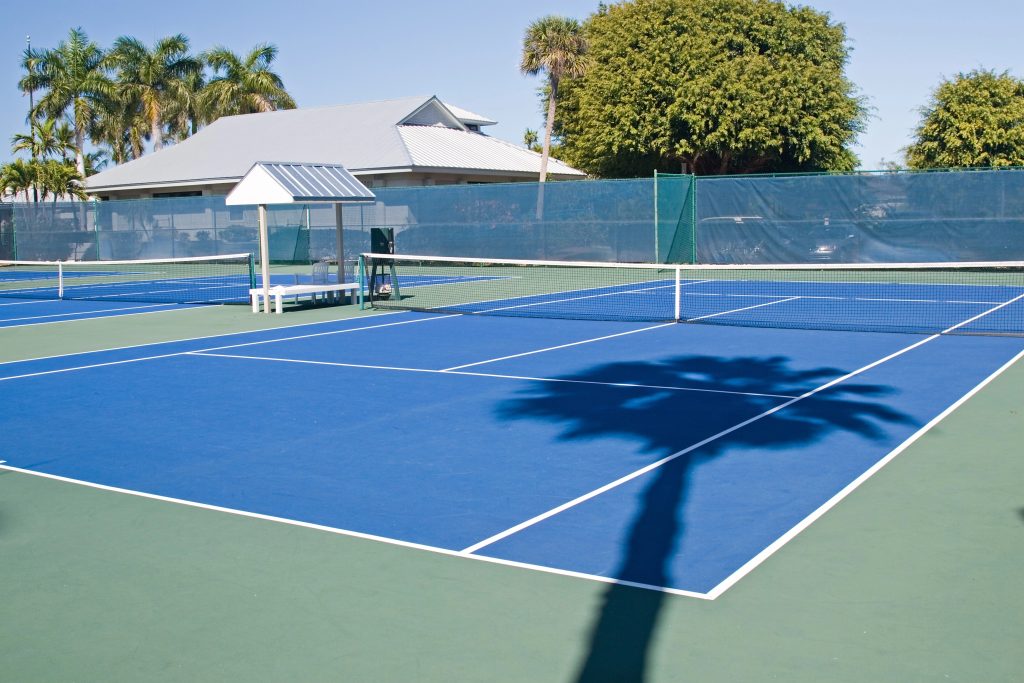 A bright blue tennis court with white lines, surrounded by green fencing and palm trees. There is a bench under a shaded area, and a large palm tree shadow is cast across the court.
