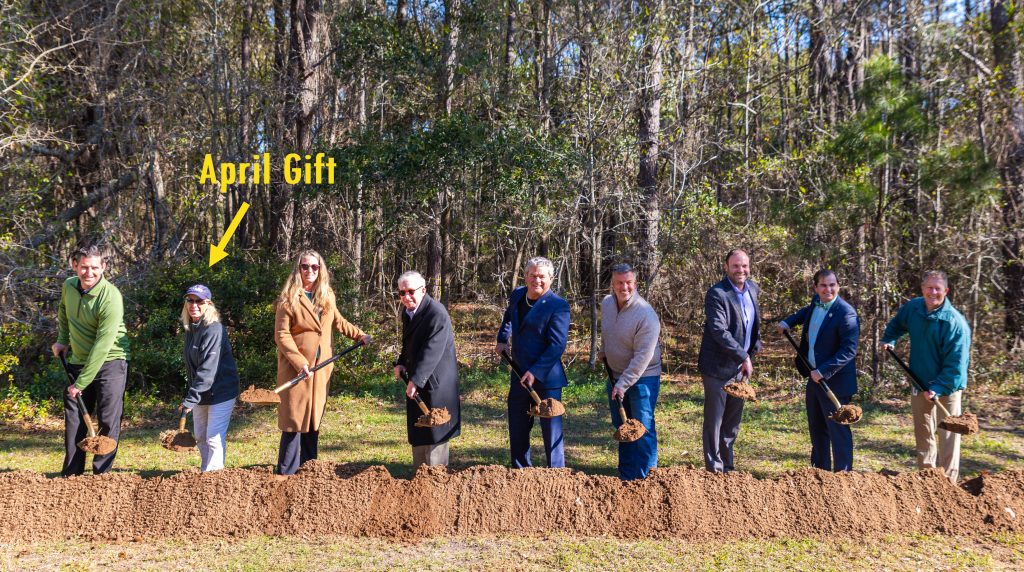 Nine people stand side by side outdoors, each holding a shovel with dirt, at a groundbreaking ceremony. A yellow arrow and the text “April Gift” point to the woman second from the left, wearing sunglasses. Trees fill the background.