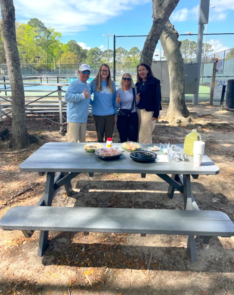 Four people stand smiling behind a gray picnic table outdoors, with food and drinks on the table. Trees and a tennis court are in the background on a sunny day.