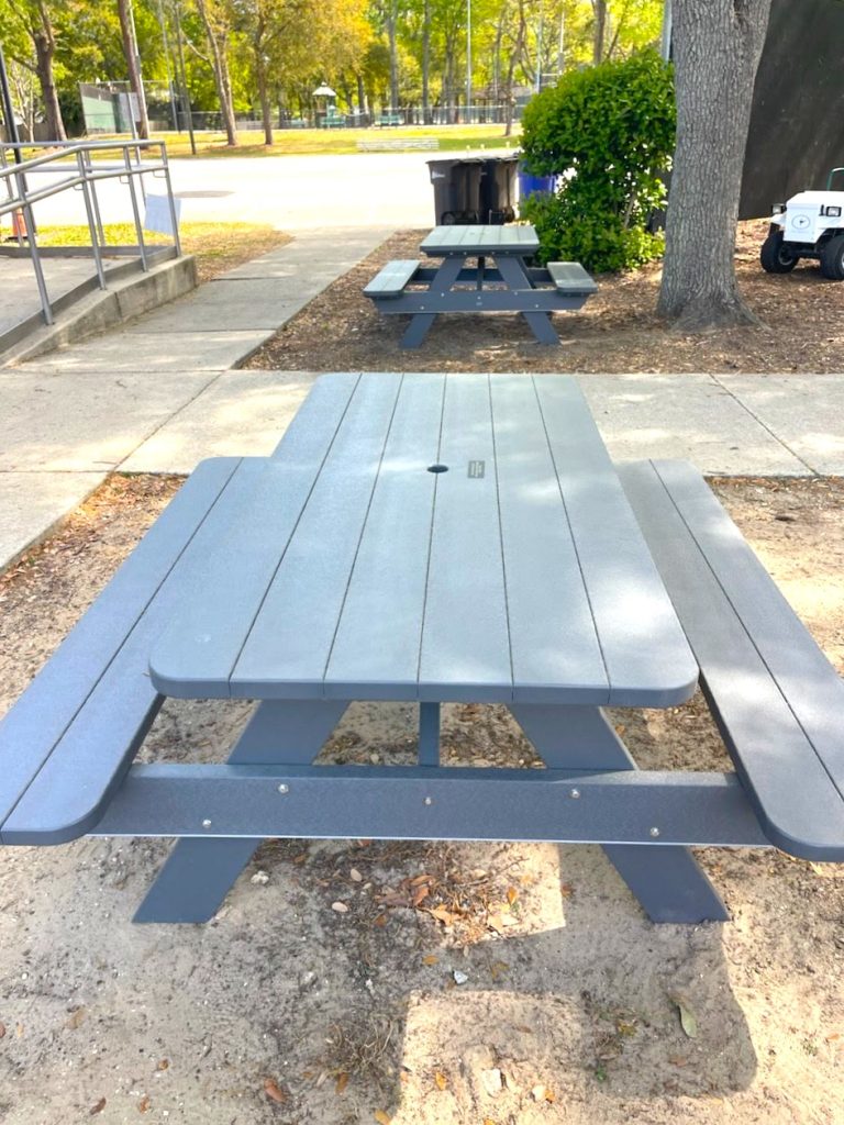 A gray picnic table sits on sandy ground near a sidewalk, with another identical table and a trash can visible in the background. Trees and green bushes surround the area.