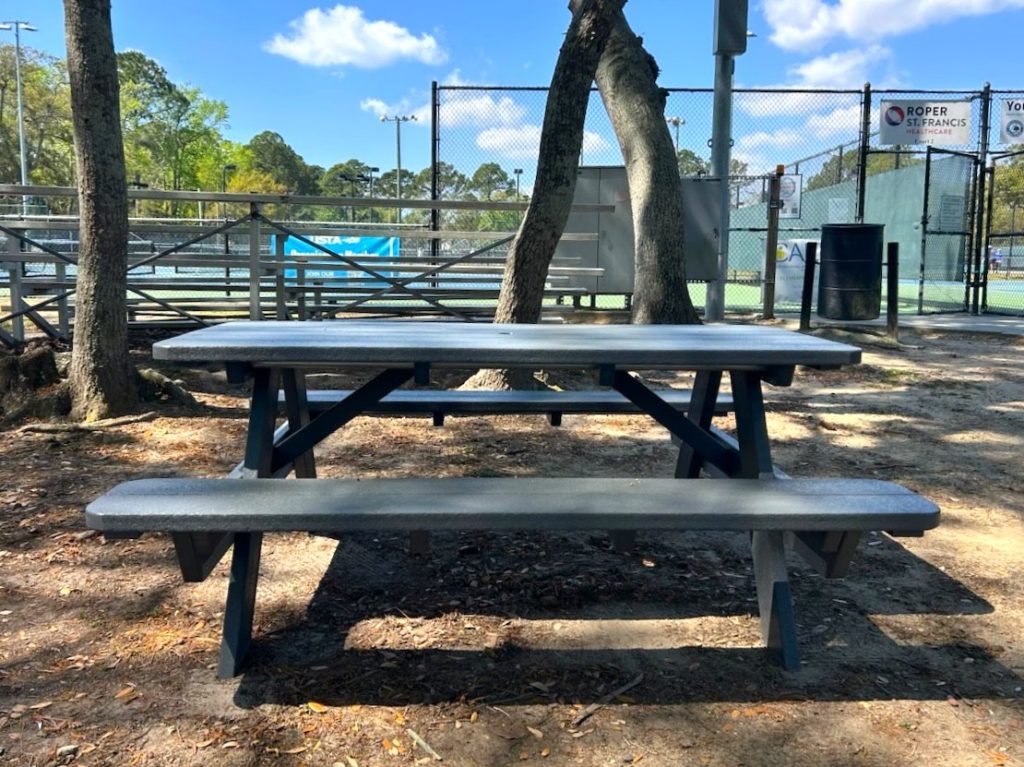 A wooden picnic table sits in the shade of trees near tennis courts, with a fence, metal bleachers, and a trash can visible in the background on a sunny day.