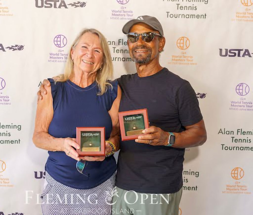 Two smiling adults, a woman and a man, stand side by side holding USTA award boxes in front of a step-and-repeat banner for the Alan Fleming Tennis Tournament.