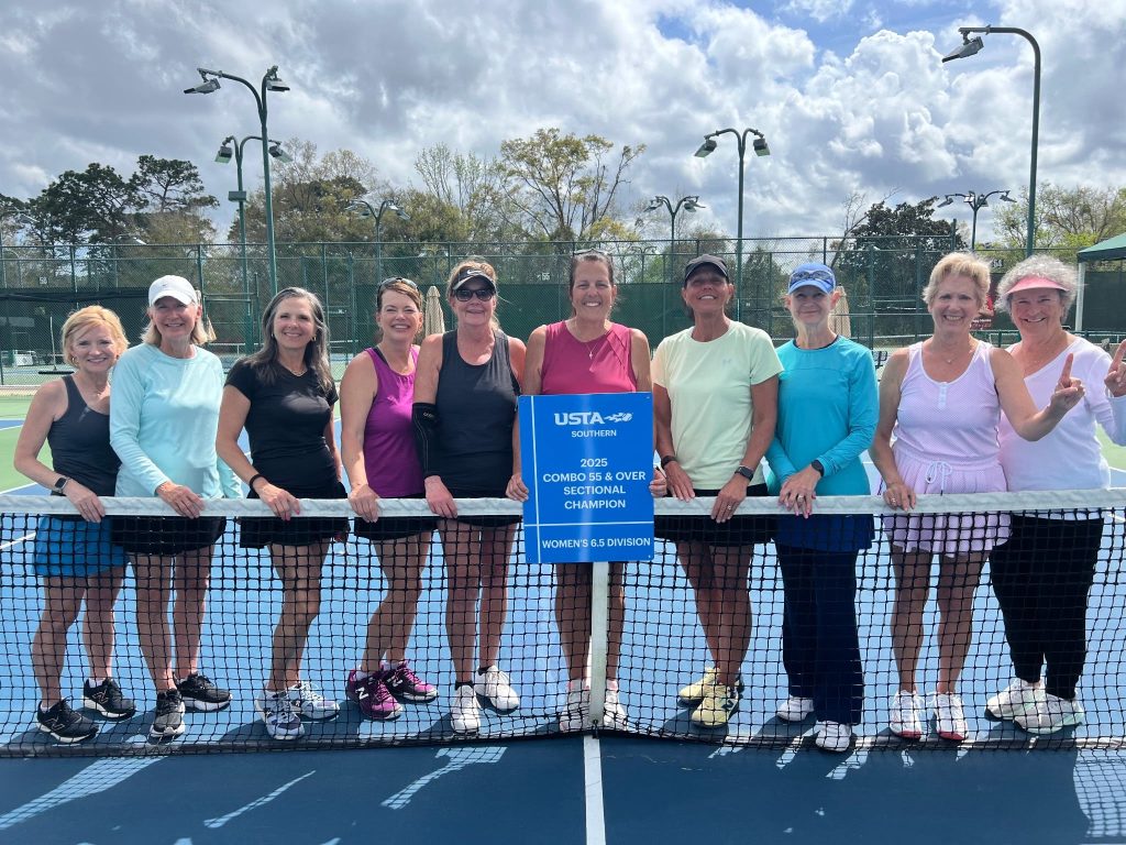 Ten women in tennis outfits stand on a tennis court behind the net, smiling and holding a blue sign that reads “USTA Southern 2023 Combo 55 & Over Sectional Champion Women’s 6.5 Division.” It is daytime with a cloudy sky.