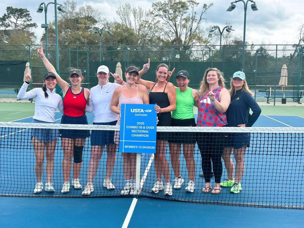 Eight women stand on a tennis court behind the net, smiling and holding a blue sign that reads