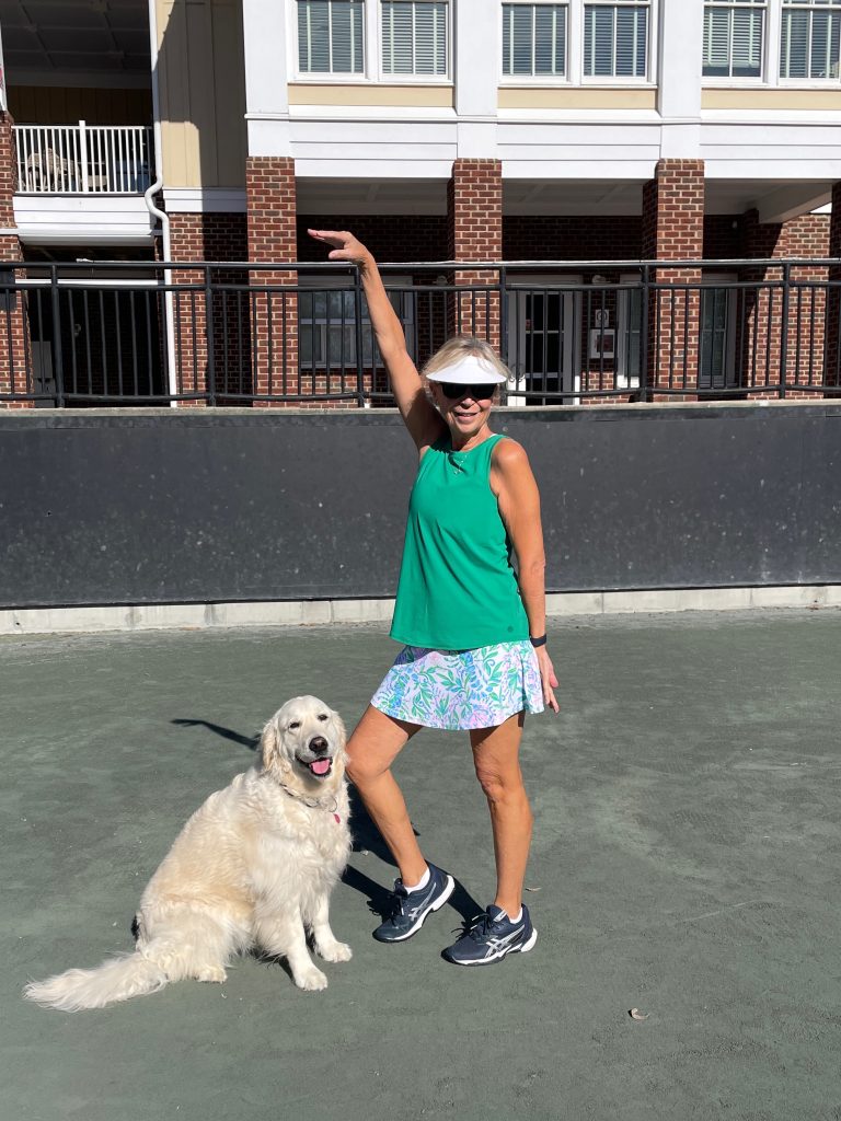 A woman in a green top, floral skirt, and sunglasses poses with one arm raised while standing on a court beside a happy, fluffy golden retriever. A brick building with white trim is in the background.