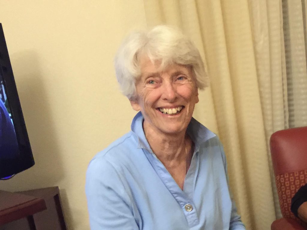 An older woman with short white hair, wearing a light blue button-up shirt, smiles warmly while sitting indoors. There is a red chair and beige curtains in the background.