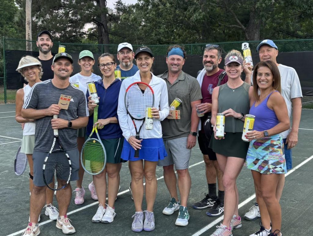 A group of thirteen smiling people stand together on a tennis court, holding tennis rackets and canned drinks. They are dressed in athletic wear, and trees and a fence are visible in the background.
