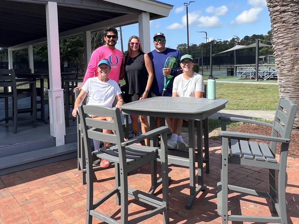 Five people are posing and smiling outdoors by a tennis court. Two are sitting at a tall patio table, while three stand behind them. It’s sunny with blue skies and scattered clouds. Tennis rackets are visible.