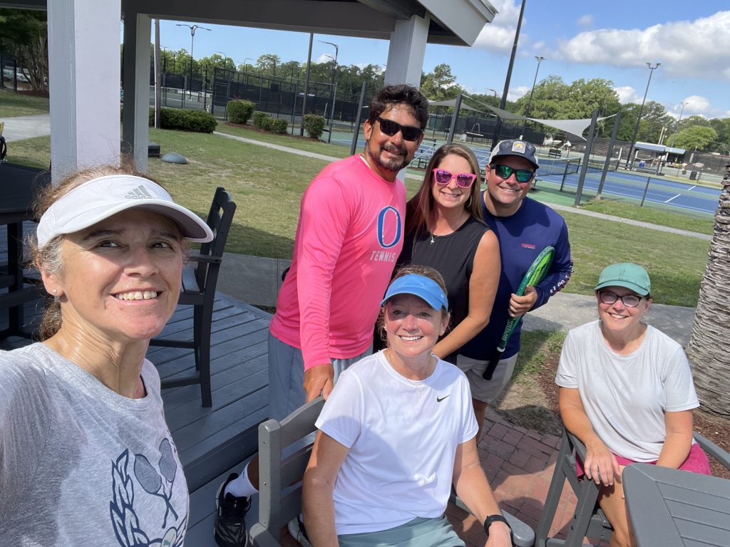Six adults, dressed in casual sportswear, smile at the camera outdoors near tennis courts on a sunny day. Some are seated at a table while others stand behind them, holding tennis gear. Trees and blue sky are in the background.