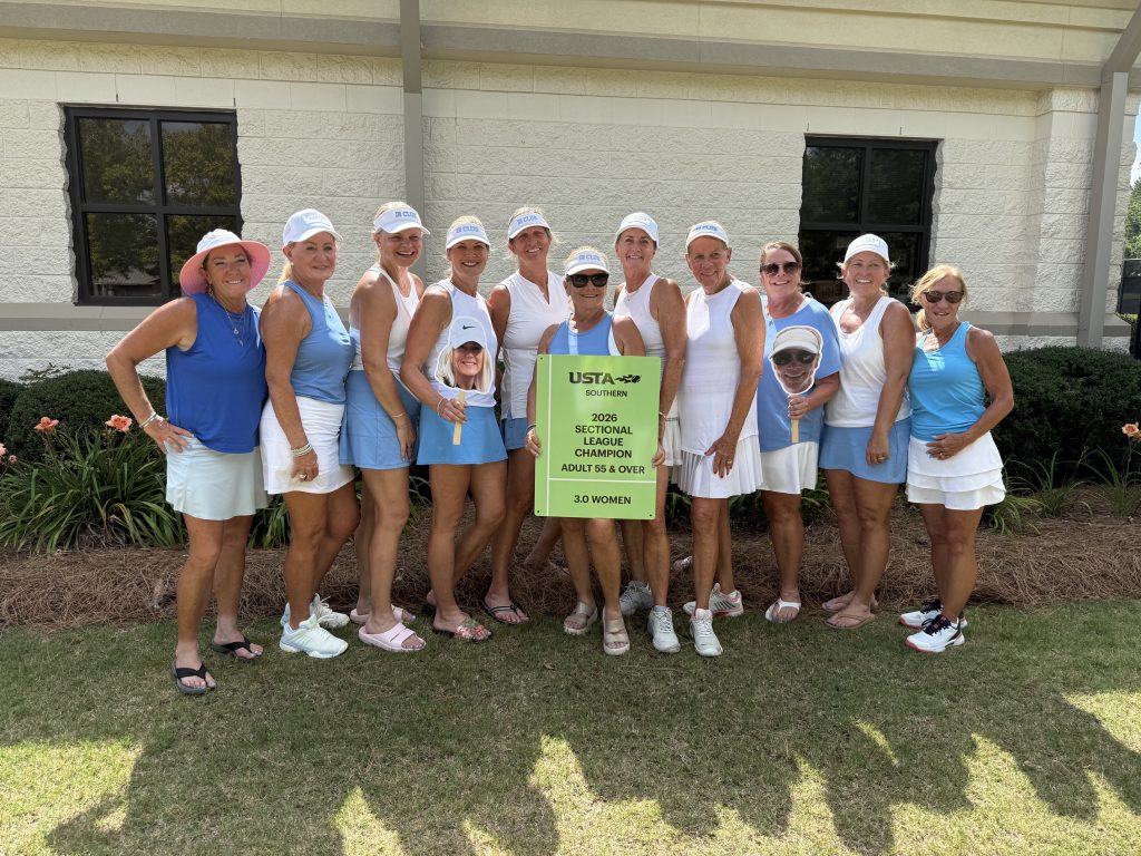 Thirteen women in tennis outfits pose outdoors, smiling and holding a green sign that reads “USTA Southern Sectional Champion Adult 55 & Over 3.0 Women,” in front of a building with flowers and bushes.