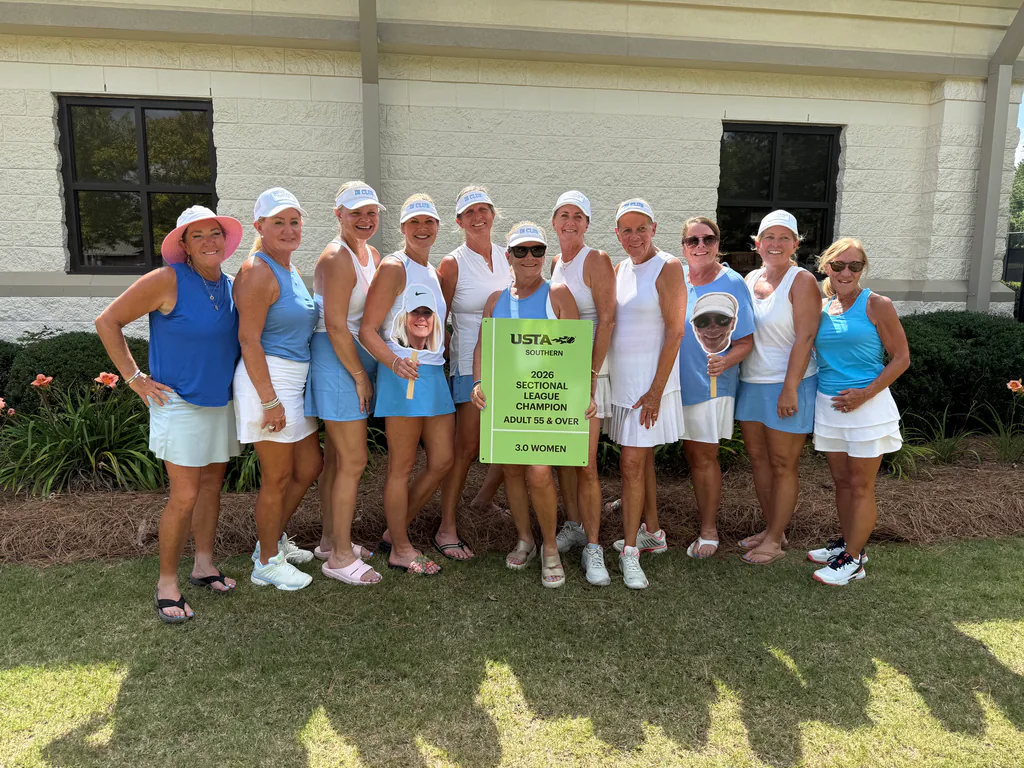 Thirteen women in tennis outfits pose outdoors, smiling and holding a green sign that reads “USTA Southern Sectional Champion Adult 55 & Over 3.0 Women,” in front of a building with flowers and bushes.