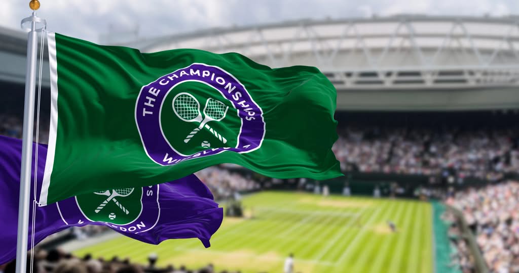 Two flags with Wimbledon logos wave in the foreground, while a blurred grass tennis court and grandstands filled with spectators are visible in the background.