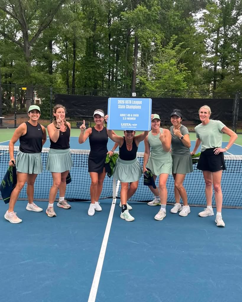 Seven women in tennis outfits stand on an outdoor tennis court, smiling and celebrating while holding a blue sign reading