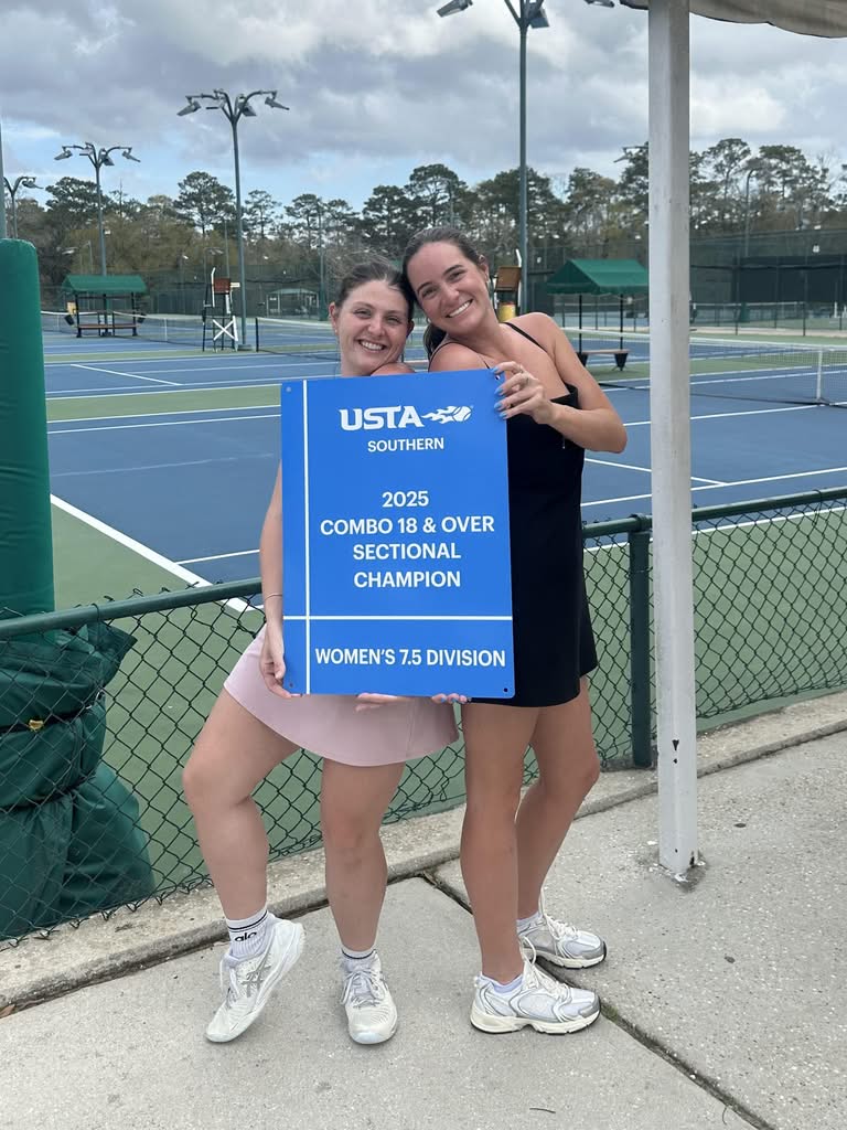 Two women in tennis outfits happily pose together by a tennis court, holding a large blue sign that reads,