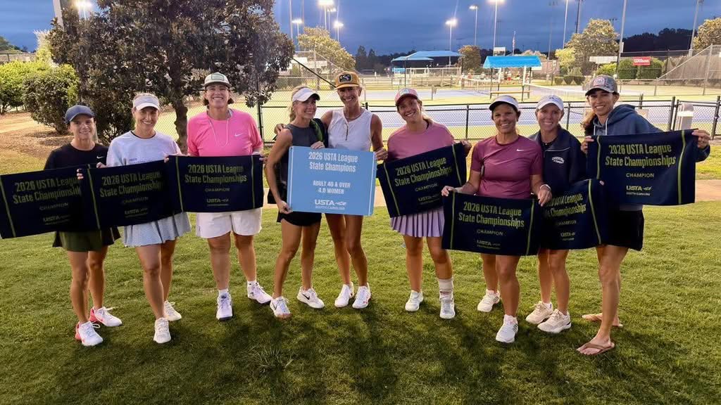 Nine women stand on grass by tennis courts, smiling and holding banners and a sign reading “2023 USTA League State Championships.” It is evening, and the tennis courts are lit in the background.