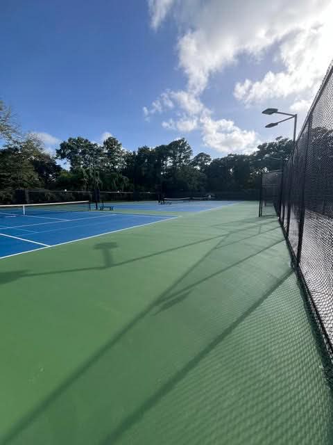 Outdoor tennis courts with blue and green surfaces, surrounded by a black chain-link fence, trees in the background, and a partly cloudy sky overhead.