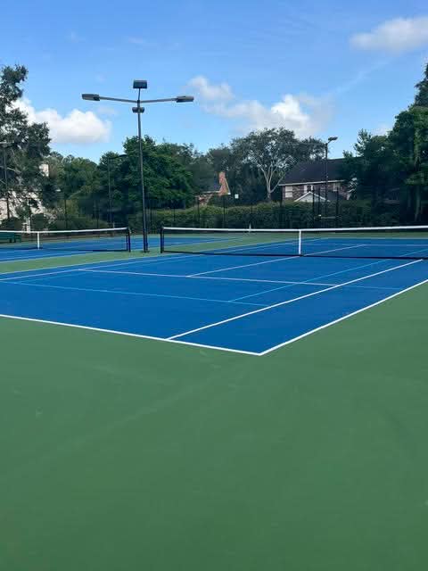 Empty outdoor tennis courts with blue playing surfaces and green outer areas, surrounded by trees, fences, and houses in the background under a sunny sky with some clouds.