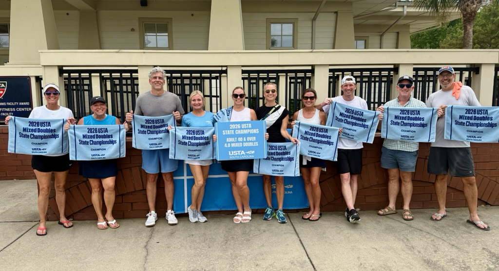 Ten adults stand in a row outdoors, each holding a blue towel that reads “2023 USTA State Mixed Doubles Champions.” They are smiling in front of a building and appear to be celebrating their victory.