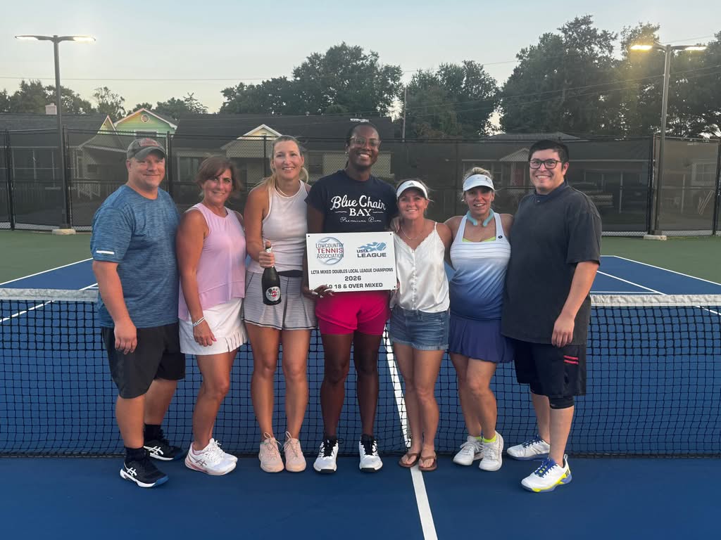 Seven adults stand smiling on a tennis court, some with arms around each other. The group holds a sign and a bottle. They appear to be celebrating a tennis league event. Trees and houses are visible in the background.