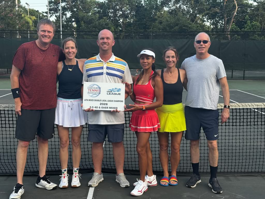 Six adults stand on a tennis court, smiling, arm in arm. Four wear tennis outfits; two wear casual clothing. They hold a sign reading “USTA League 2023 40 & Over Mixed Local Champion.” Trees and a fence are in the background.