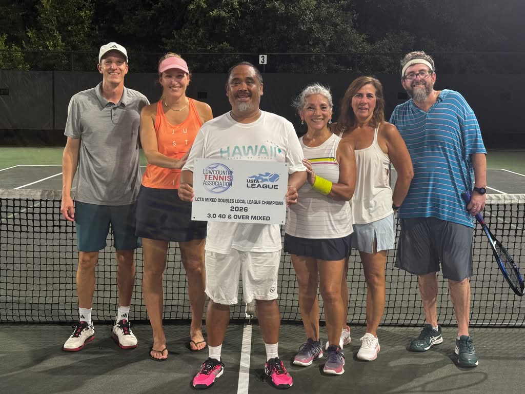 Six adults stand on a tennis court at night, smiling and holding a sign that reads “USTA Mixed Doubles League Champions 2023. 3.0 40 & Over Mixed.” They wear casual tennis attire and stand in front of a net.