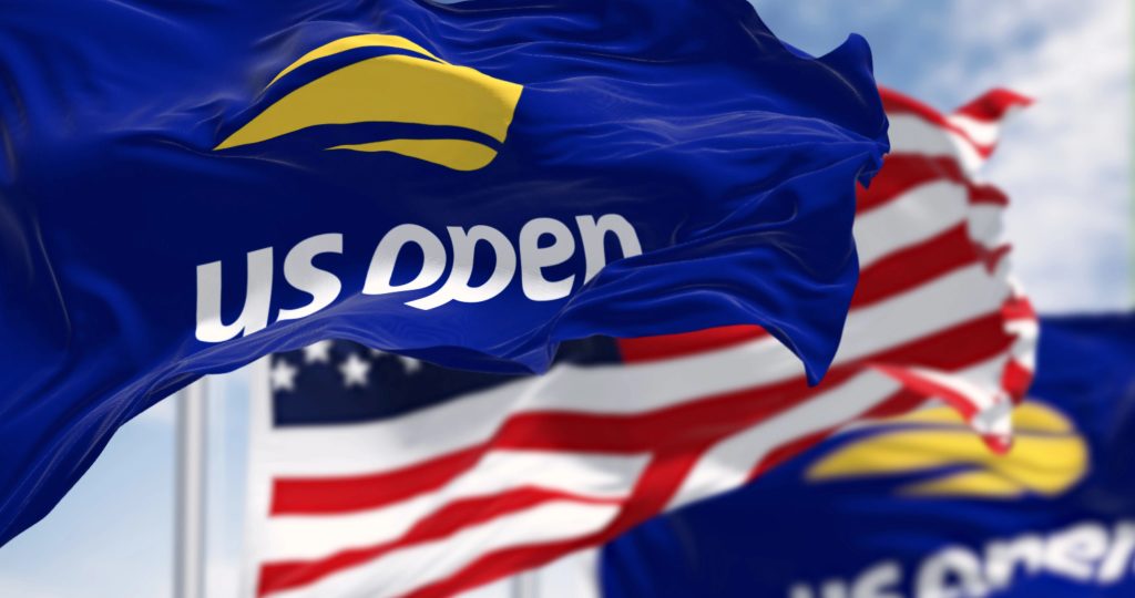 Blue US Open flags with yellow tennis ball logo wave in the foreground, while American flags fly in the background against a partly cloudy sky.