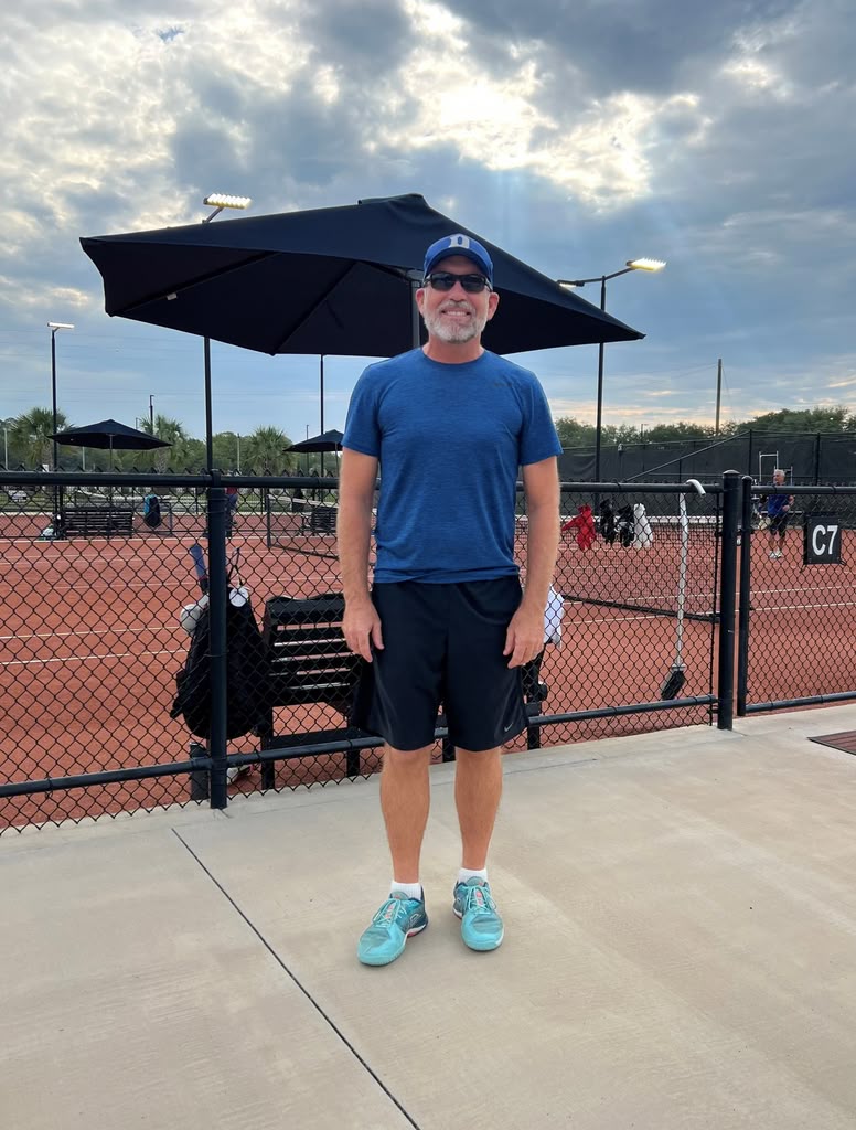 A man in blue sportswear and sunglasses stands on a tennis court sidewalk, smiling. Tennis courts, benches, and large black umbrellas are visible in the background under a cloudy sky.