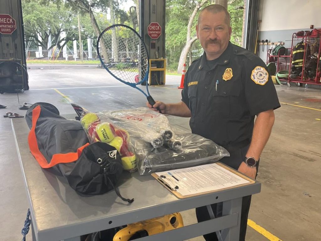 A uniformed firefighter stands in a fire station beside a table holding rescue equipment and a clipboard, smiling at the camera.