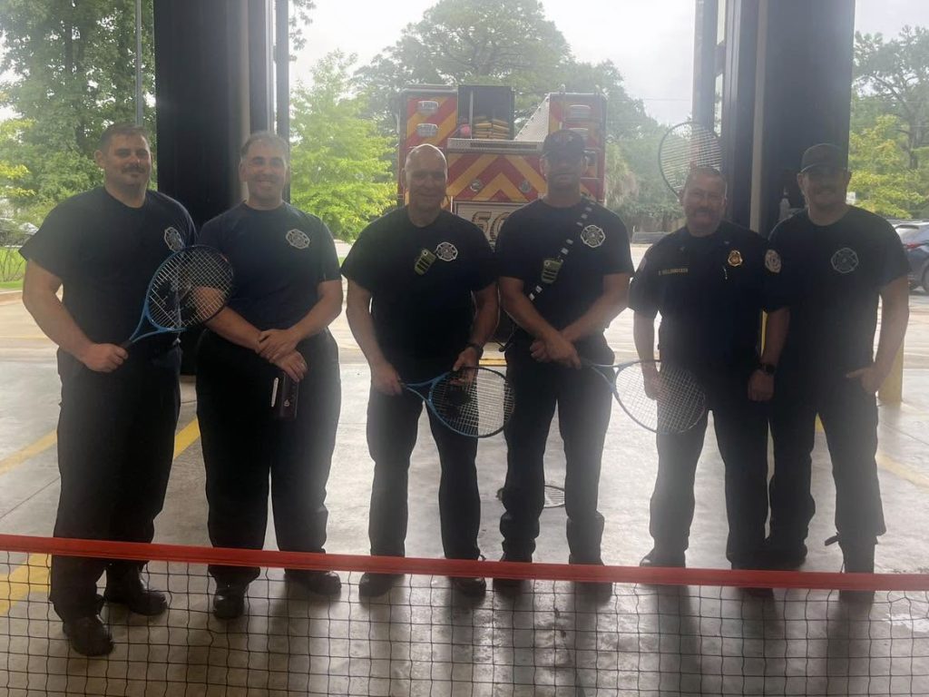 Six firefighters in uniform stand indoors holding pickleball paddles behind a net, with a fire truck and trees visible in the background.