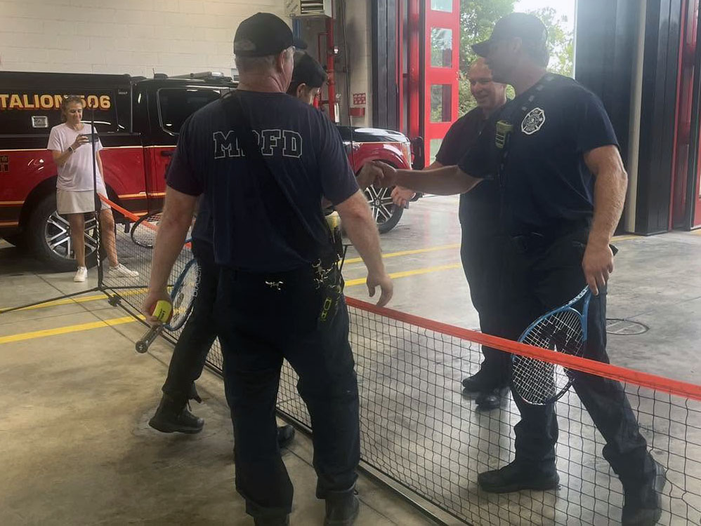 Four firefighters shake hands over a pickleball net inside a fire station, holding paddles. A woman in the background watches and a red fire vehicle is visible outside.