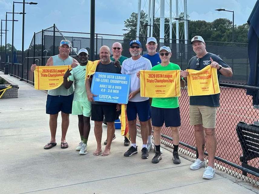 Eight men stand on a tennis court holding championship towels and a banner that reads “2023 USTA League 7.0 Level Champions Adult 55 & Over.” They are smiling, posing for a group photo in front of a chain-link fence.