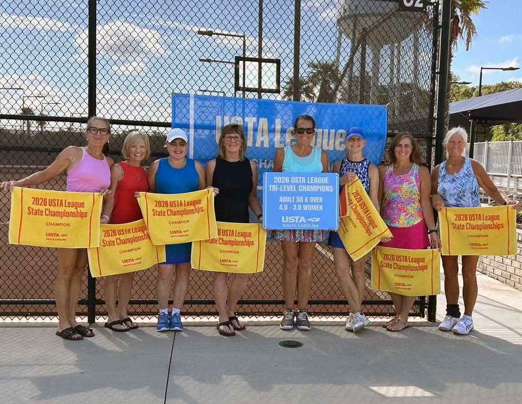 Eight women stand in front of a tennis court holding 2024 USTA League State Championships banners and a sign for 2024 USTA League Tri-Level Champions. They are smiling and dressed in tennis attire.