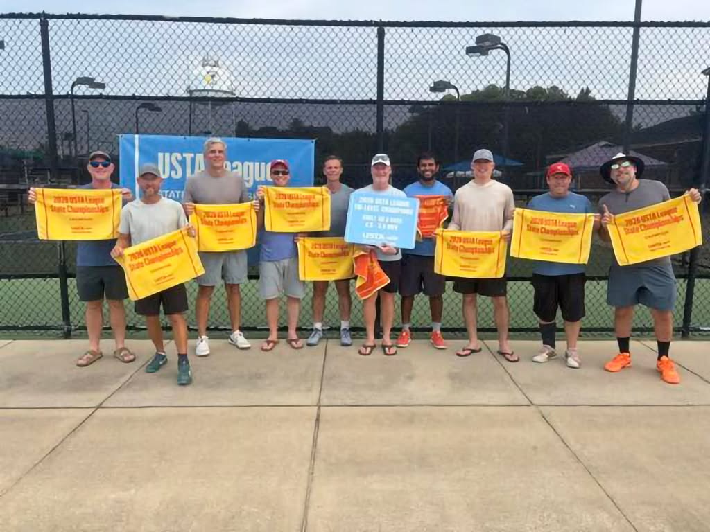 Ten people stand on a tennis court holding bright yellow “USTA Tennis League Champions” towels, smiling for a group photo. A fence and blue banner are visible in the background.