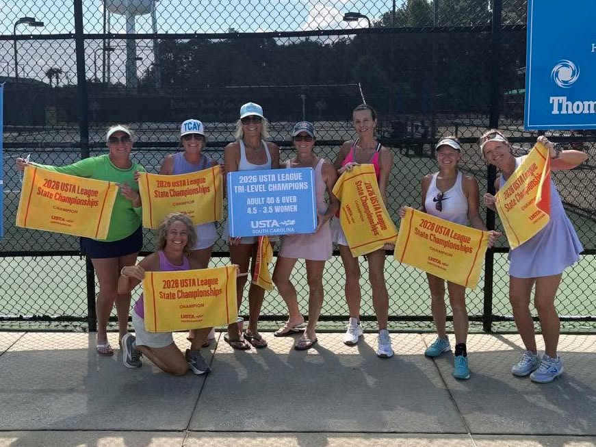 Eight women pose on a tennis court holding yellow and red 2023 USTA League State Championship towels and a blue 2023 USTA Tri-Level Champions banner, smiling on a sunny day.