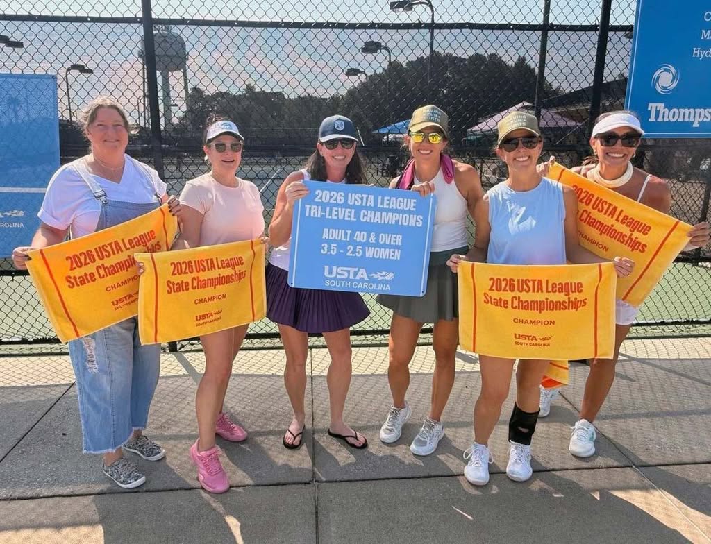 Six women stand on a tennis court holding orange towels and a blue sign that reads “2026 USTA League Tri-Level Champions Adult 40 & Over 3.5-2.5 Women.” They are smiling and dressed in tennis outfits.