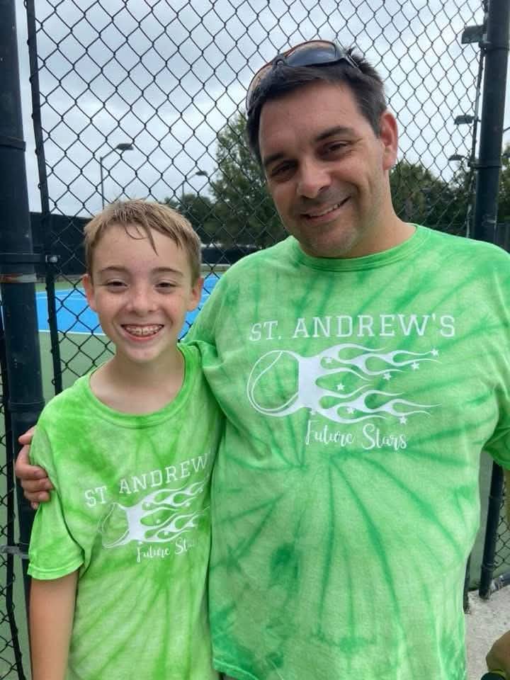 A smiling man and boy in matching green “St. Andrew’s Future Stars” tennis shirts stand together by a fence on a cloudy day.